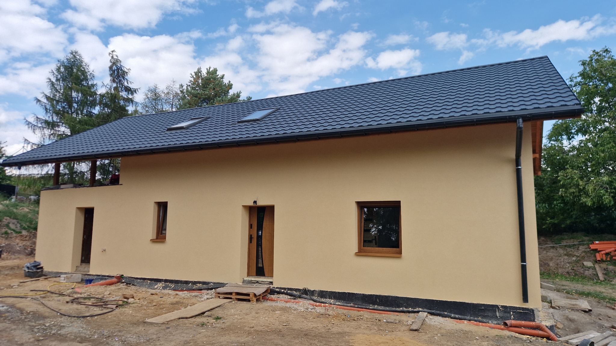 Nowoczesny dom jednorodzinny z beżową elewacją i ciemnym, metalowym dachem. Widoczne okna dachowe i elementy wykończeniowe. Wokół domu teren w trakcie prac ziemnych.