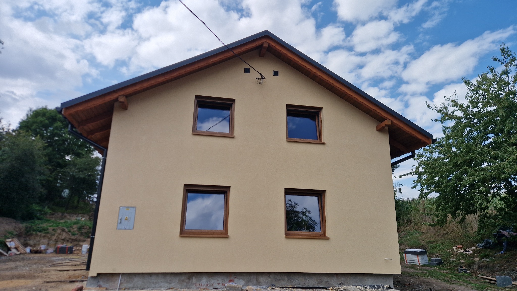 Dwukondygnacyjny dom z beżową elewacją i drewnianymi ramami okiennymi w słoneczny dzień z widocznymi elementami wykończeniowymi dachu i instalacją elektryczną.