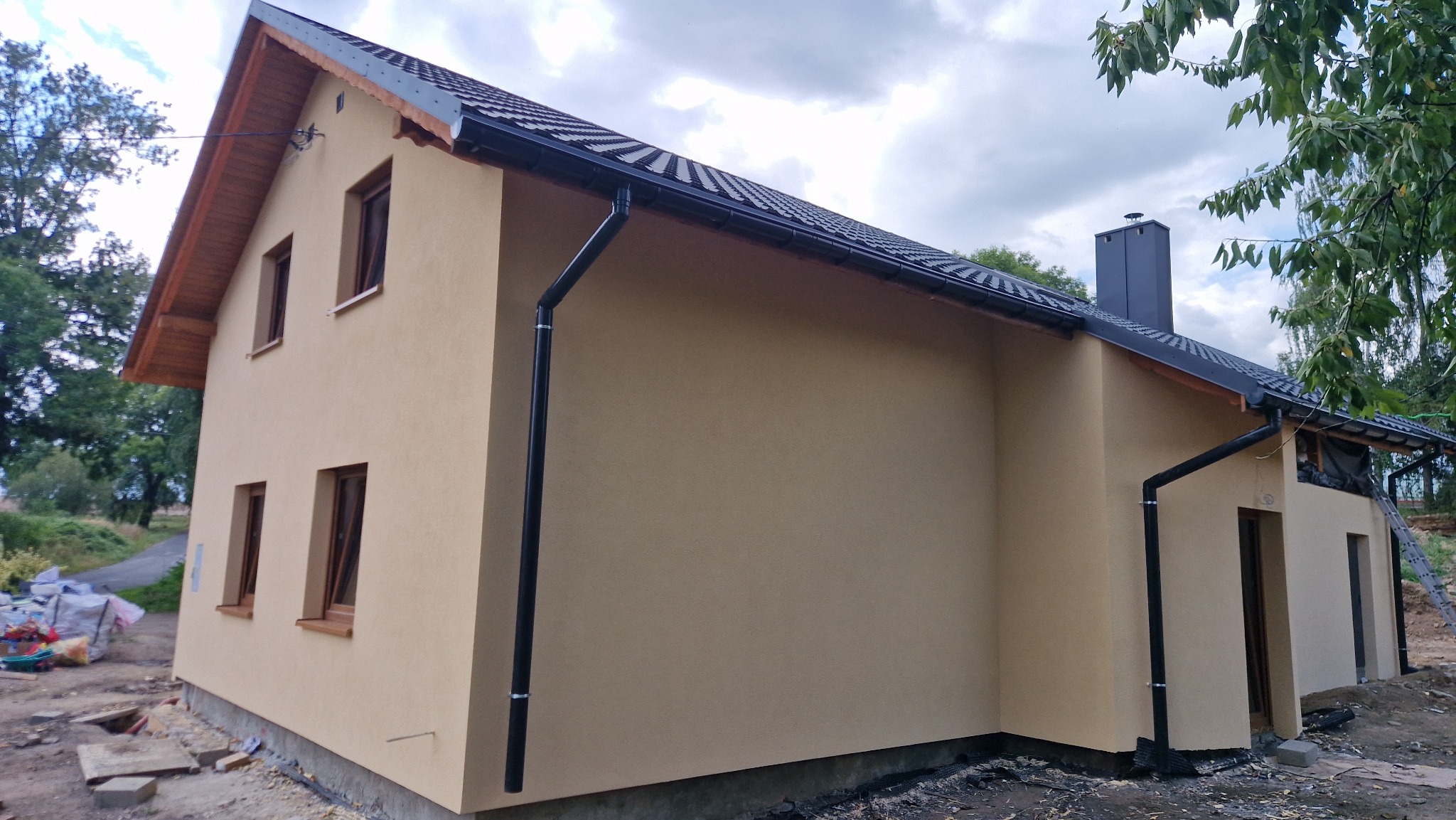 Dwukondygnacyjny dom z beżową elewacją i ciemnymi rynnami, widoczny z boku. Dach pokryty czarną dachówką. Okna z brązowymi ramami. Częściowo widoczne drzewa w tle.