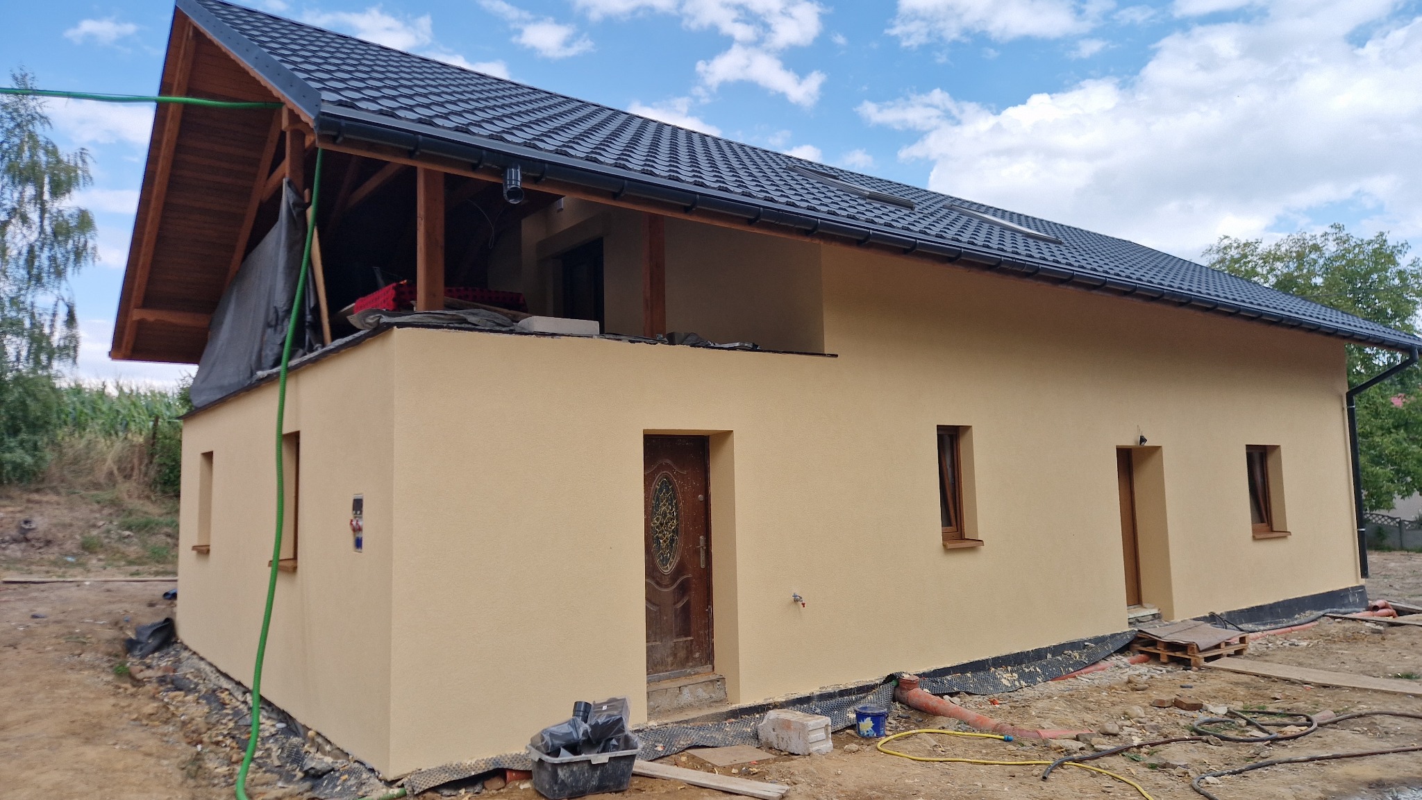 Niski dom z beżową elewacją i ciemnym dachem w trakcie budowy. Widoczne drewniane elementy konstrukcyjne i narzędzia budowlane. Częściowo wykończona elewacja.