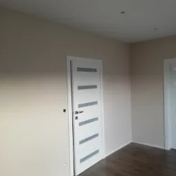 Jasne pomieszczenie po remoncie, z beżowymi ścianami, białymi drzwiami z szarymi akcentami i podłogą z ciemnego drewna, widoczne gniazdko elektryczne bez osłony.