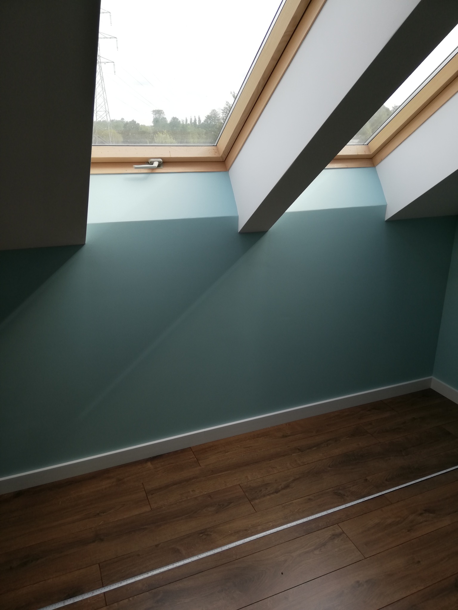 Ściana w odcieniu turkusu z oknami dachowymi i drewnianą podłogą, widoczny cień padający ze skosu dachu, listwa przypodłogowa w kolorze białym.