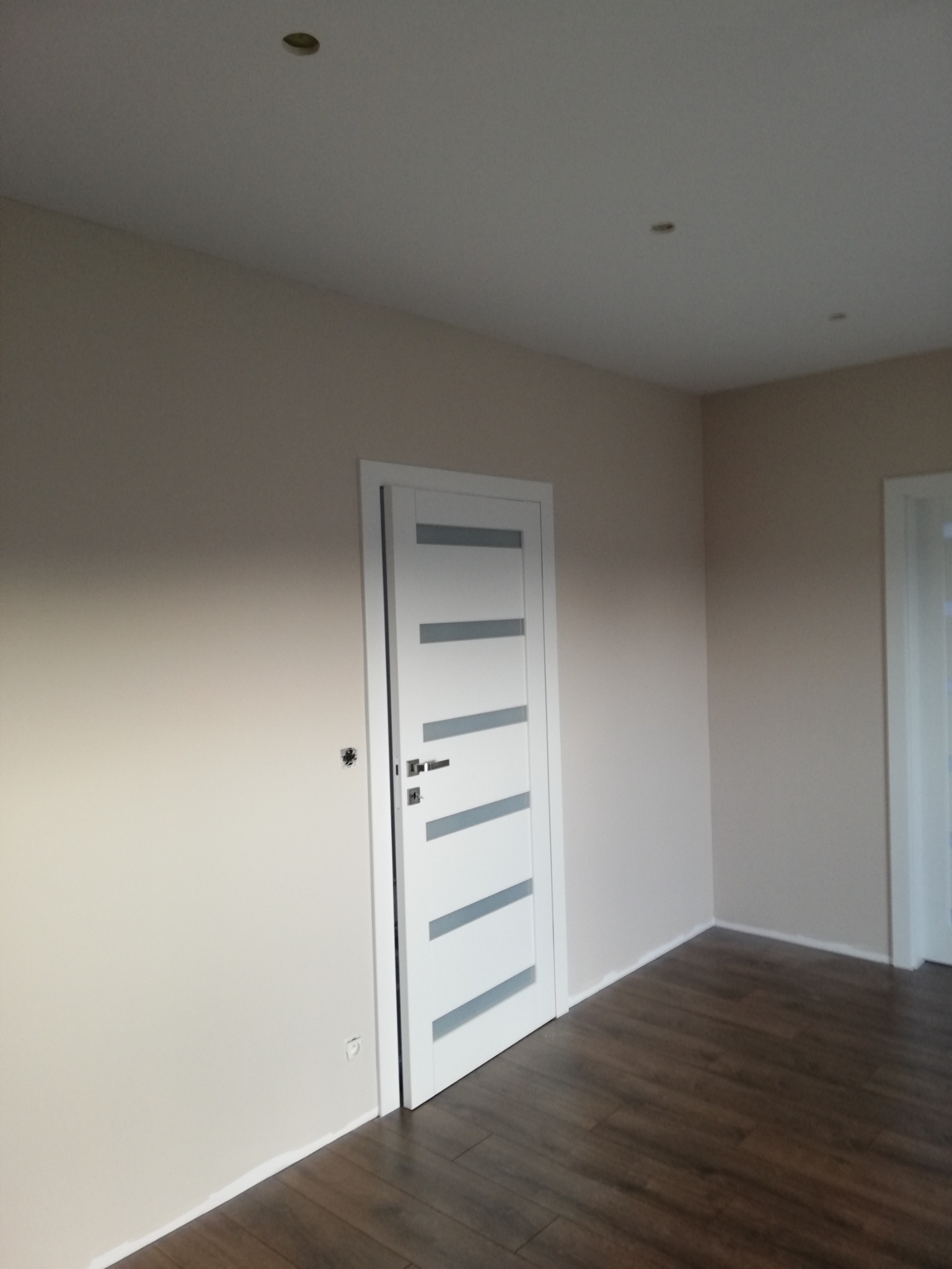 Jasne pomieszczenie po remoncie, z beżowymi ścianami, białymi drzwiami z szarymi akcentami i podłogą z ciemnego drewna, widoczne gniazdko elektryczne bez osłony.