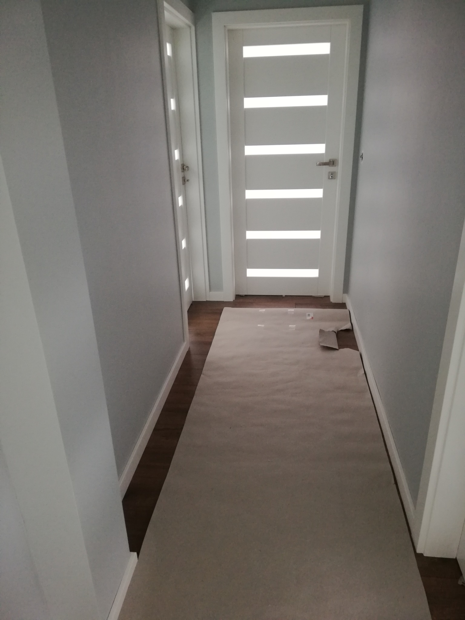Korytarz zabezpieczony papierem malarskim przed rozpoczęciem prac, ściany w kolorze jasnoszarym, białe drzwi z przeszkleniami.