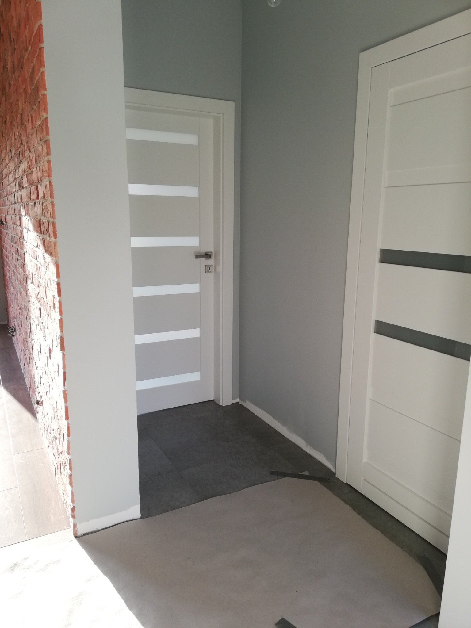 Korytarz po odświeżeniu z szarą ścianą, białą stolarką drzwiową z przeszkleniami i fragmentem ściany z cegły, zabezpieczony podłogą folią malarską.