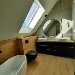 Nowoczesna łazienka na poddaszu z drewnopodobnymi płytkami, owalną wanną wolnostojącą, czarną armaturą i okrągłym lustrem pod oknem dachowym.