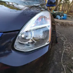 Błyszczący reflektor i fragment karoserii ciemnego SUV-a, z odbiciami drzew na lakierze, na tle nieuporządkowanego otoczenia z workami i sprzętem.