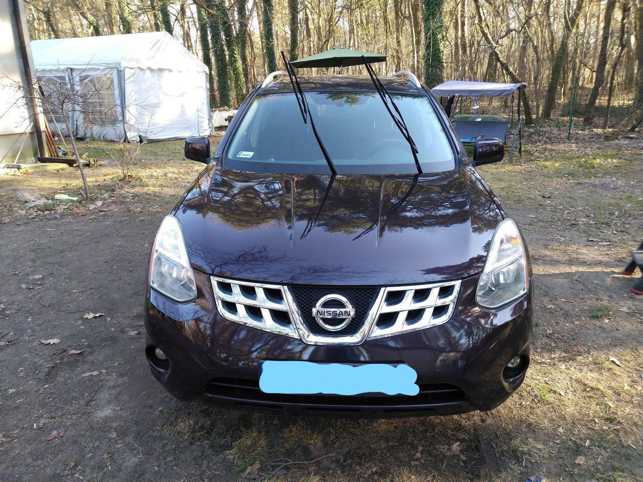 Ciemny Nissan Rogue przed domem w Warce, z małym parasolem na wycieraczkach, sugerujący mycie lub suszenie. W tle widoczny namiot i huśtawka ogrodowa.
