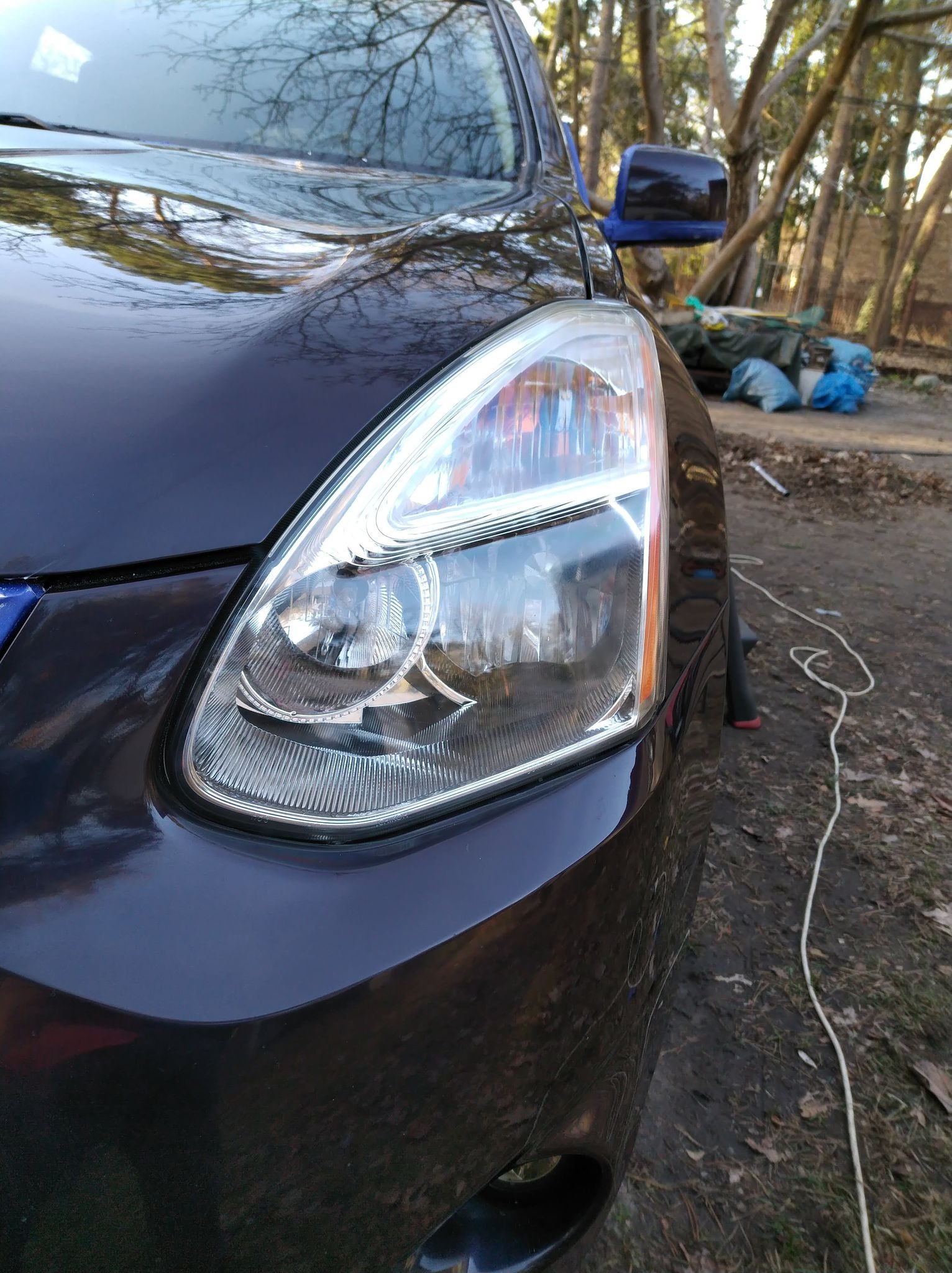 Błyszczący reflektor i fragment karoserii ciemnego SUV-a, z odbiciami drzew na lakierze, na tle nieuporządkowanego otoczenia z workami i sprzętem.