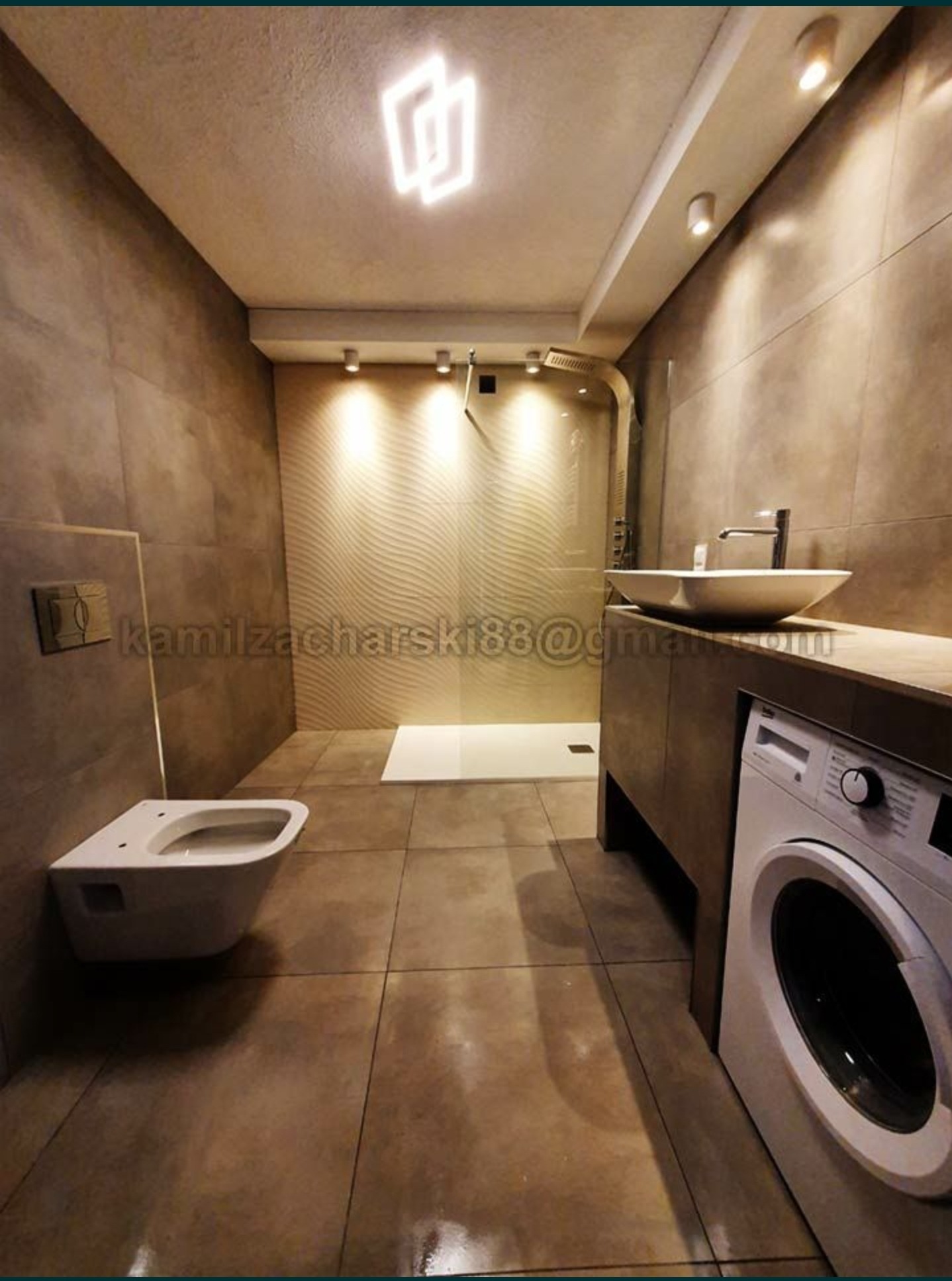 Nowoczesna łazienka z beżowymi płytkami, podwieszaną toaletą, umywalką nablatową i pralką, oświetlona geometryczną lampą sufitową i punktowymi reflektorami.