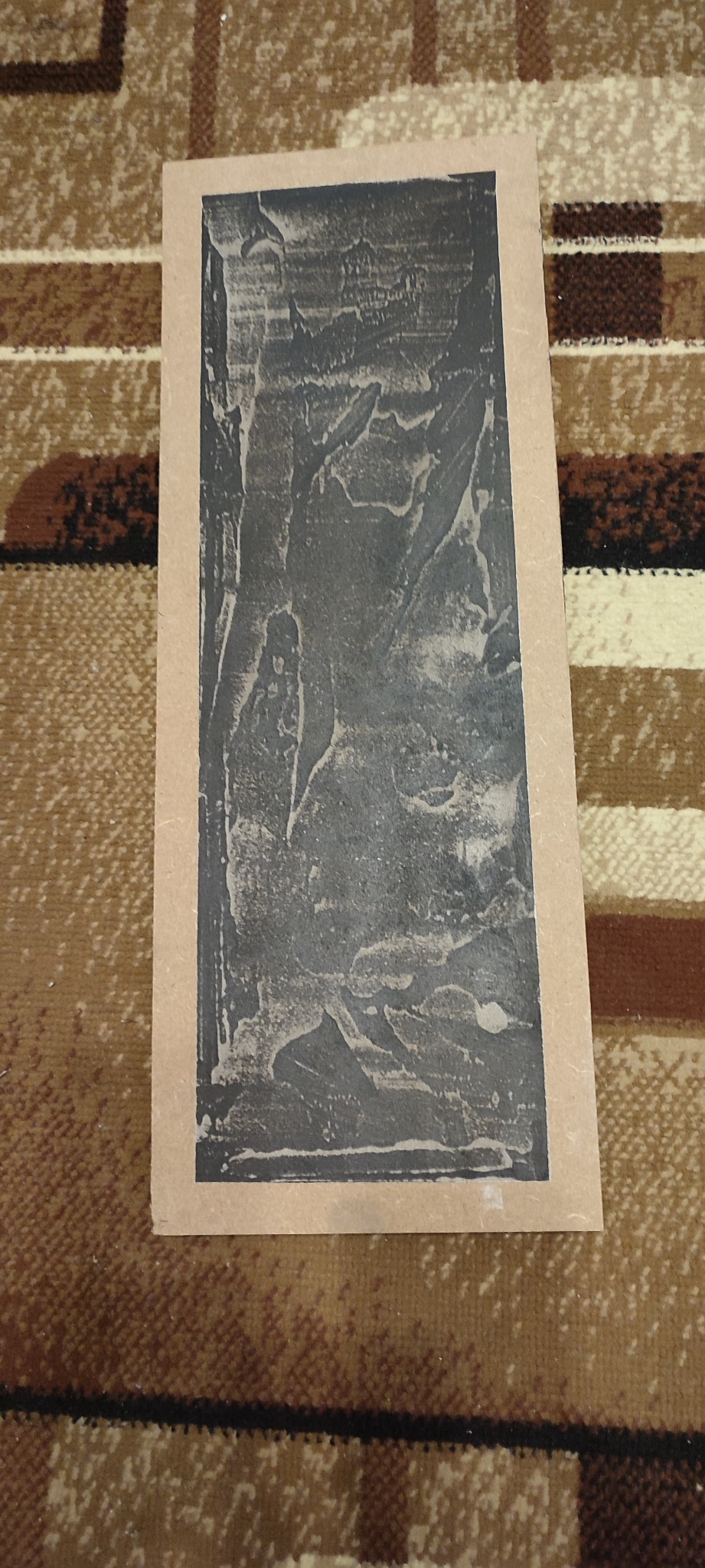 Próbka faktury szpachli w odcieniach szarości na kartonowym podkładzie, leżąca na dywanie z geometrycznym wzorem.