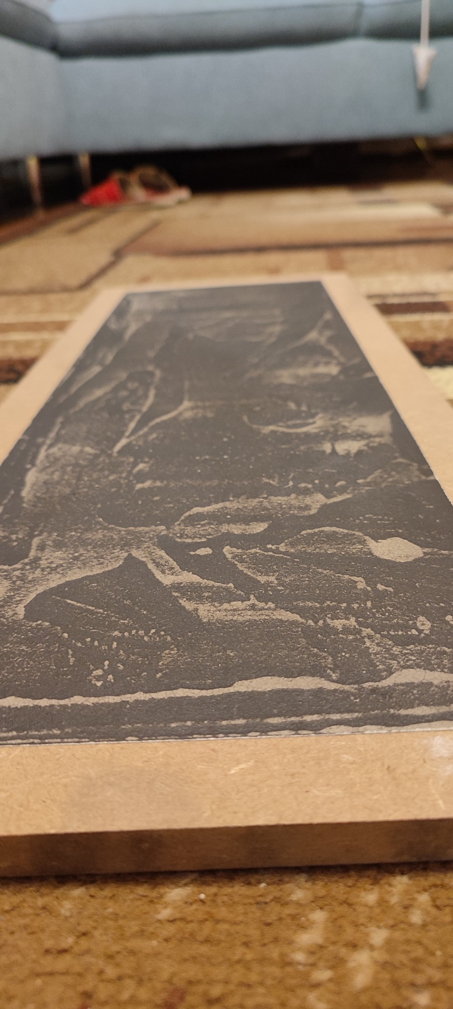 Nisko kątowe ujęcie płyty gipsowo-kartonowej ze śladami szpachlowania, umieszczonej na podłodze z widocznym wzorem dywanu i fragmentem sofy w tle.