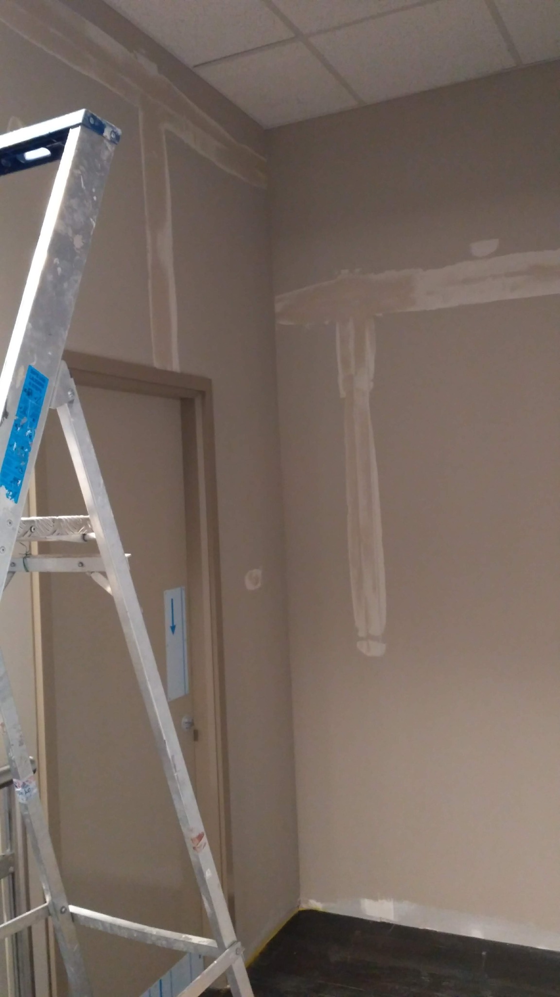 Fragment wnętrza w trakcie przygotowania ścian do malowania, widoczna drabina aluminiowa, pionowe i poziome pasy świeżo nałożonej masy szpachlowej na ścianach, sufit kasetonowy.