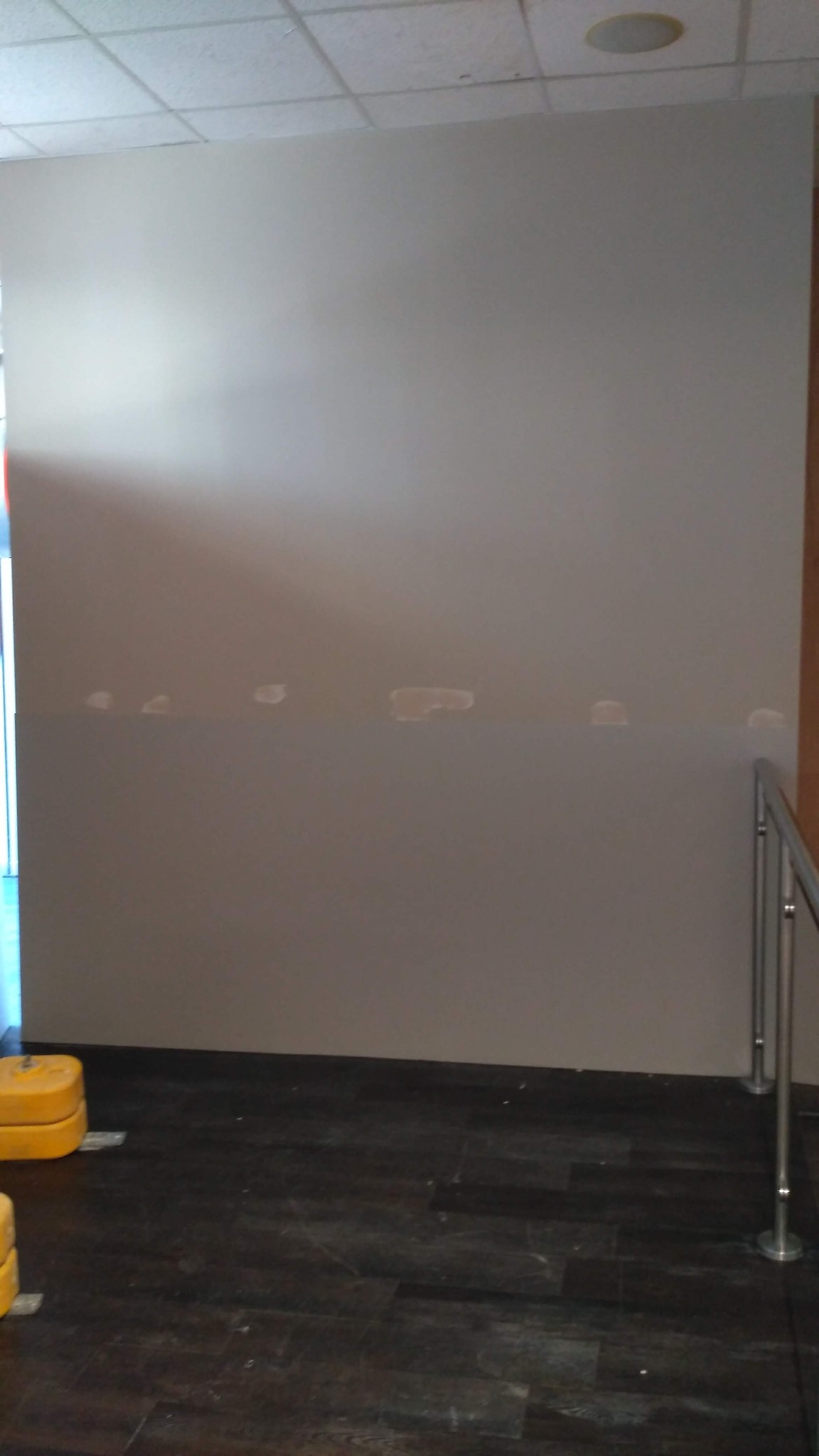 Szara ściana w trakcie przygotowania do malowania, z widocznymi miejscami po szpachlowaniu, ciemna podłoga i fragment metalowej barierki.