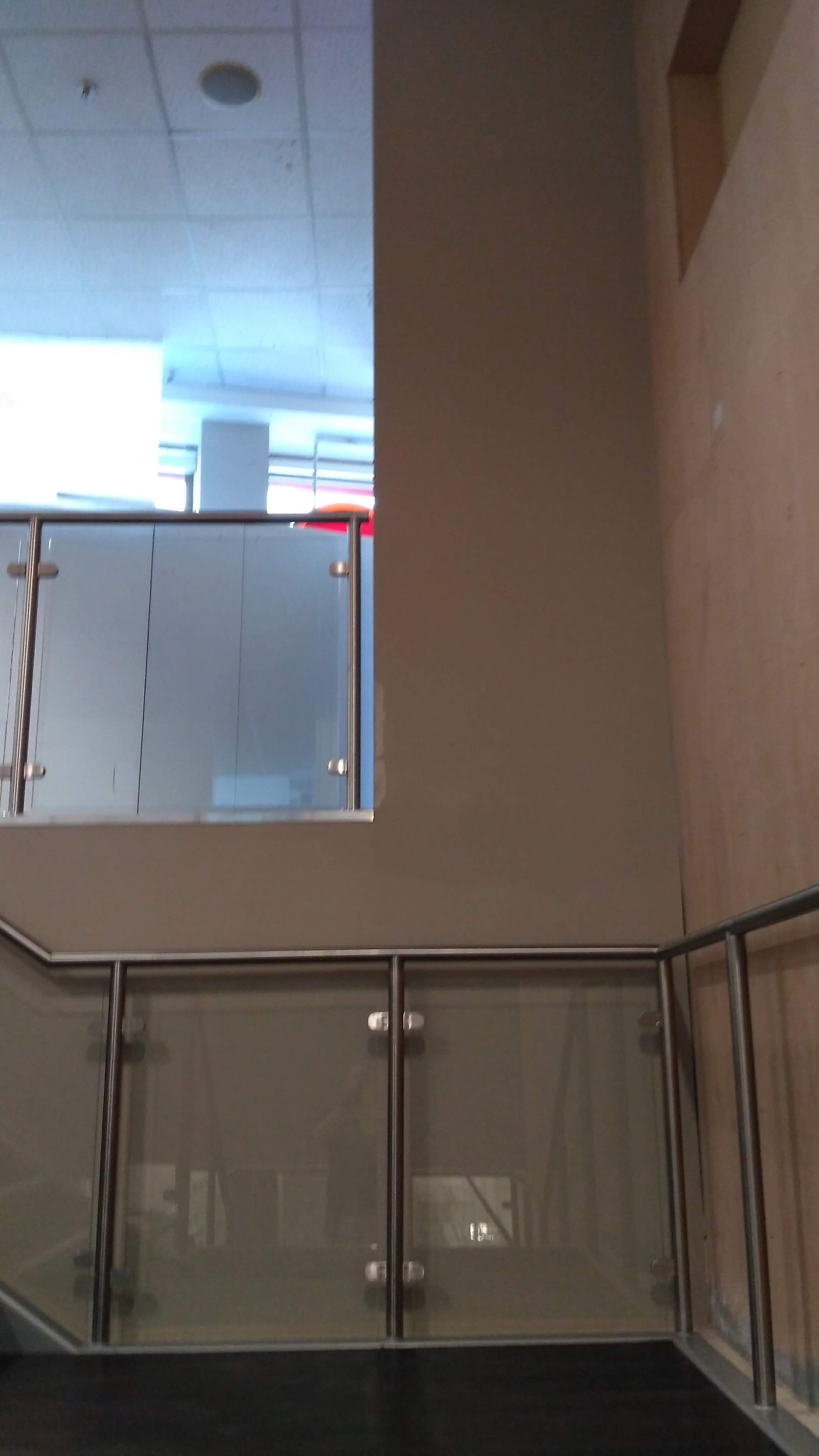 Wnętrze biura z przeszkloną balustradą na schodach, widoczna ściana pomalowana na beżowo i sufit z oświetleniem rastrowym.