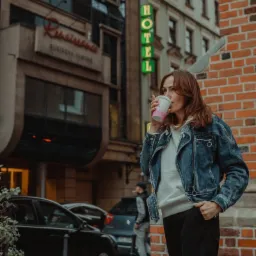 Kobieta z brązowymi włosami w dżinsowej kurtce pije kawę na tle ceglanej ściany i budynku z neonem Hotel.
