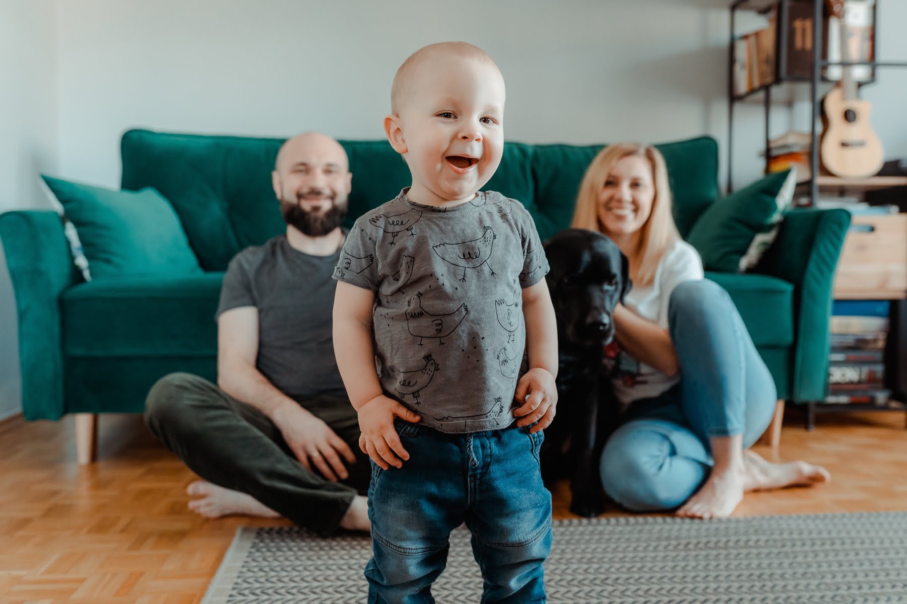 Uśmiechnięty roczny chłopiec w koszulce z kurczakami stoi na dywanie, w tle rodzice i czarny labrador siedzą na podłodze przed szmaragdową sofą.
