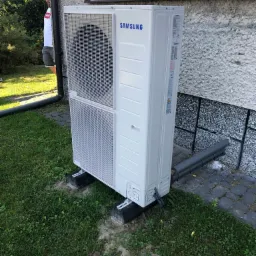 Biała jednostka zewnętrzna z logo Samsung, zamontowana na betonowych podstawach przy ścianie domu z widoczną izolacją rur i fragmentem trawnika.