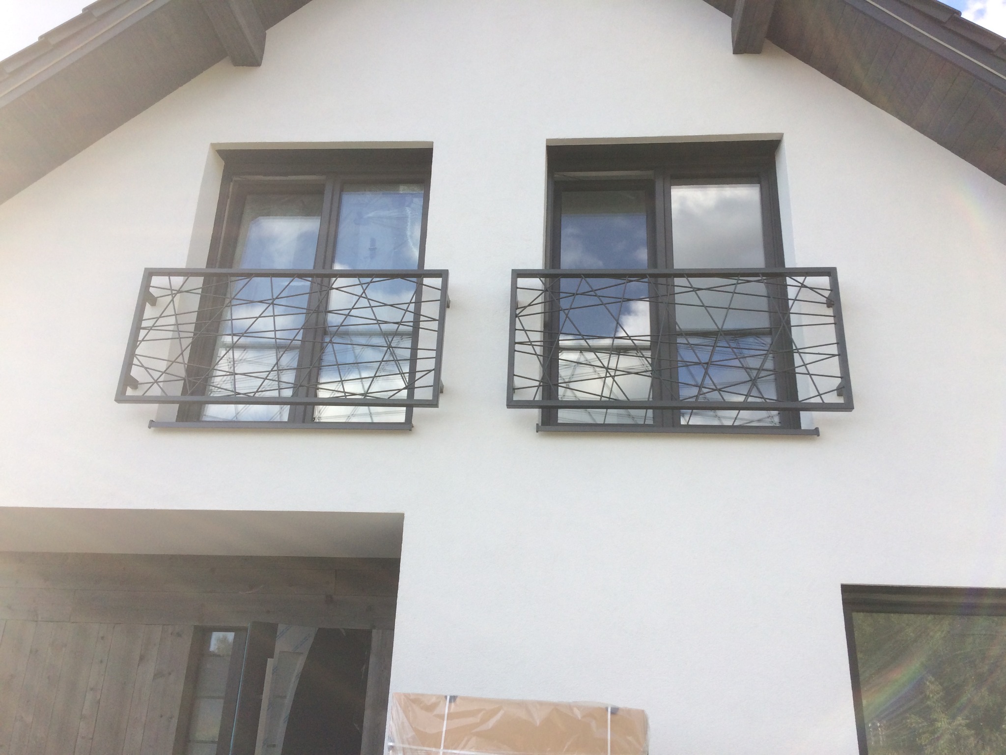Nowoczesna fasada domu z dwoma oknami zabezpieczonymi metalowymi balustradami o geometrycznym wzorze, odbijającymi błękitne niebo.