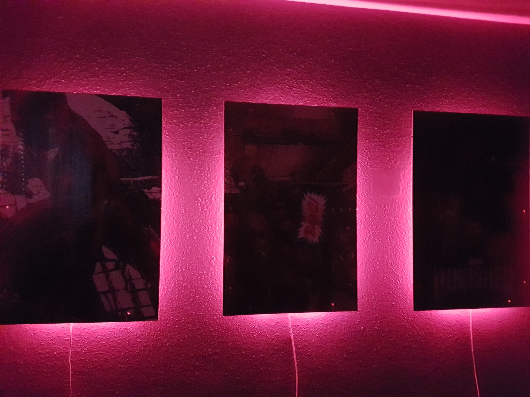 Trzy podświetlone plakaty na teksturowanej ścianie, oświetlone różowym światłem z góry i dołu, z widocznymi kablami zasilającymi.