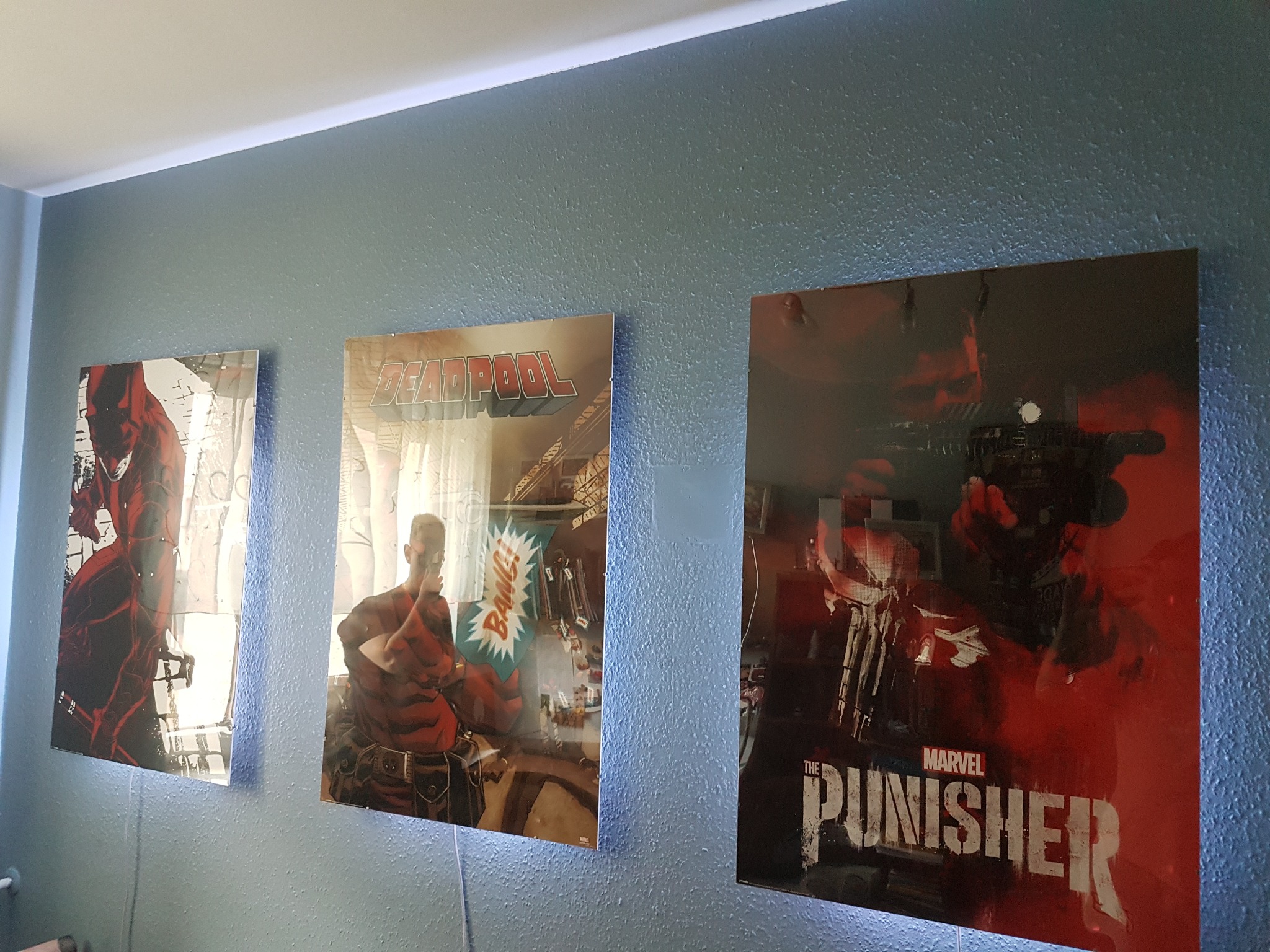 Trzy podświetlane panele LED z grafikami Daredevila, Deadpoola i Punishera zawieszone na niebieskiej ścianie z widoczną fakturą, oświetlone od góry listwą LED.