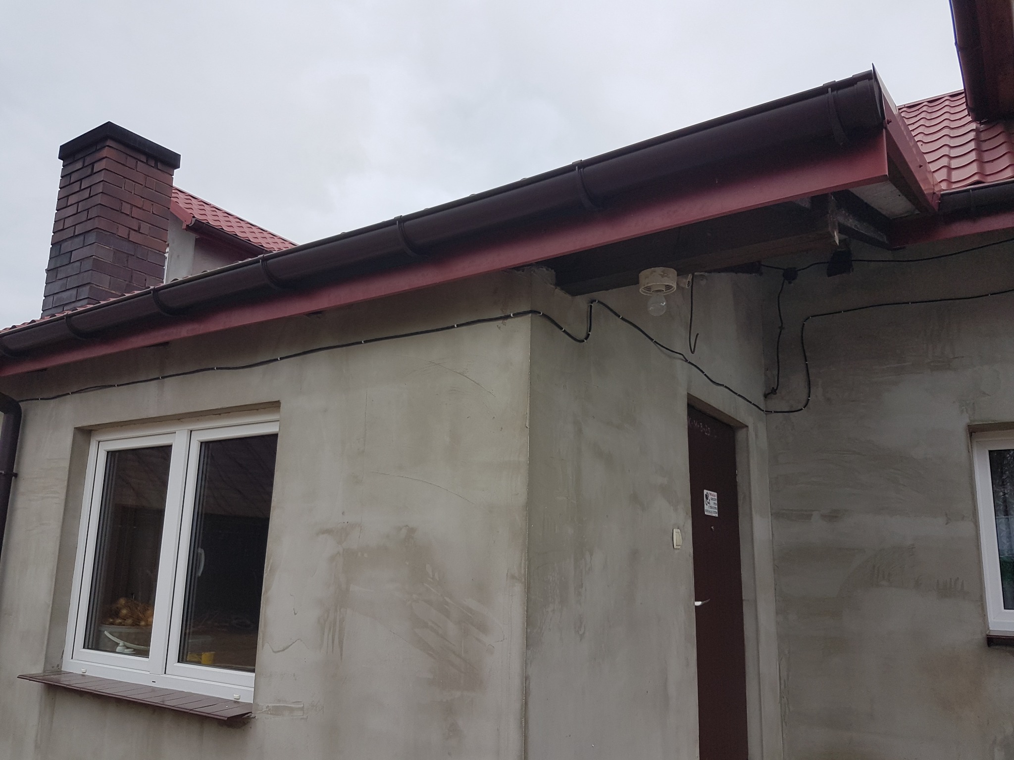 Surowy budynek w trakcie wykończenia elewacji, widoczne okno z parapetem, drzwi wejściowe z tabliczką informacyjną, zamontowane orynnowanie w kolorze brązowym oraz wystające przewody elektryczne...