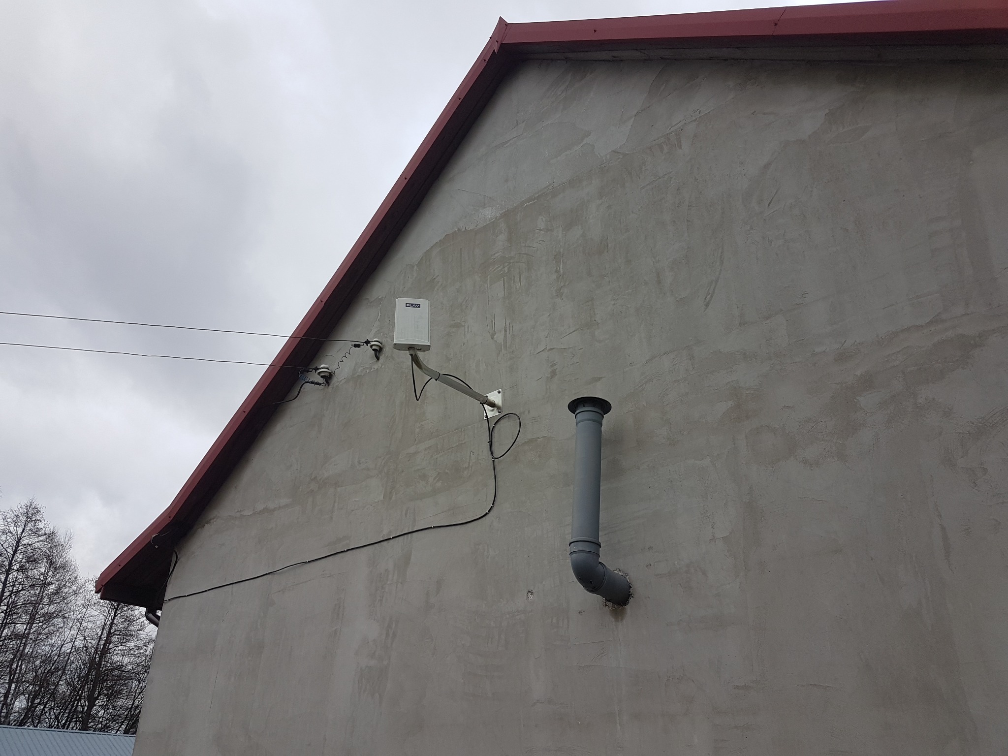 Boczna ściana budynku z szarym tynkiem, widoczna puszka instalacyjna z anteną, kabel elektryczny i szara rura wentylacyjna.