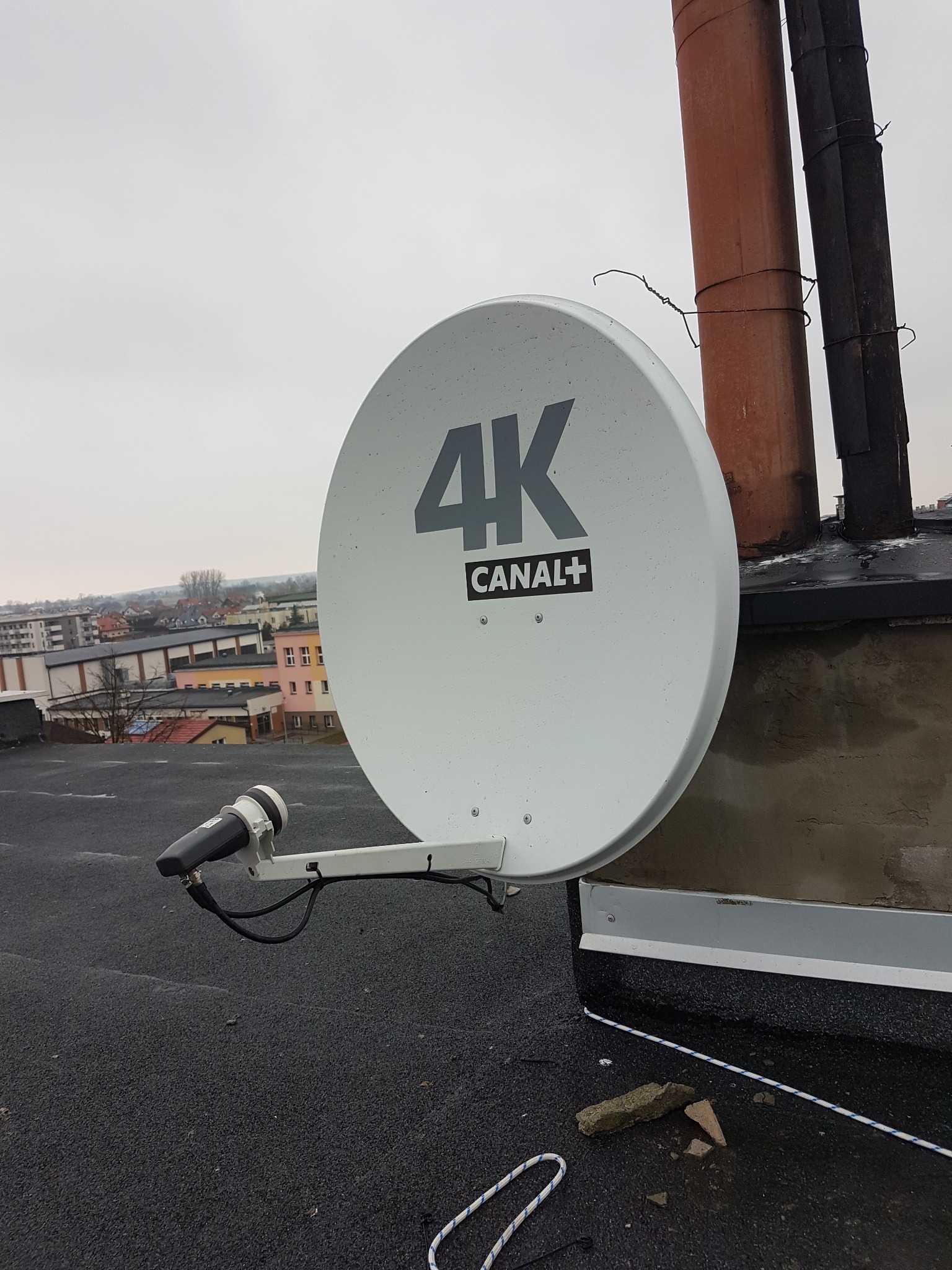Biała antena satelitarna 4K Canal+ zamontowana na płaskim dachu budynku, z widocznymi kominami i zabudową miejską w tle, sfotografowana w pochmurny dzień.