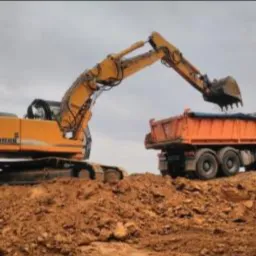 Żółta koparka Liebherr ładuje ziemię do pomarańczowej ciężarówki na tle pochmurnego nieba.