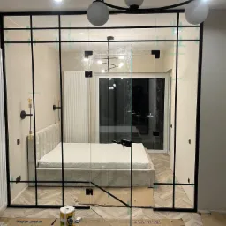 Szklana ścianka działowa w stylu loft z czarnymi profilami, oddzielająca sypialnię, widoczne taśmy zabezpieczające na szkle, kartony na podłodze sugerują trwający montaż.