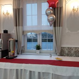 Firma Usługowa Finezja - Stół z białym obrusem, czerwonym bieżnikiem, dekoracjami balonowymi, automatem do kawy i podgrzewaczem wody, ustawiony przy oknie z widokiem na zewnątrz, ozdobiony rośliną doniczkową.
