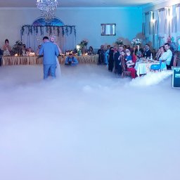 Firma Usługowa Finezja - Para młoda tańczy pierwszy taniec w dymie na sali weselnej, goście siedzą przy stołach, widoczny sprzęt do wytwarzania dymu.