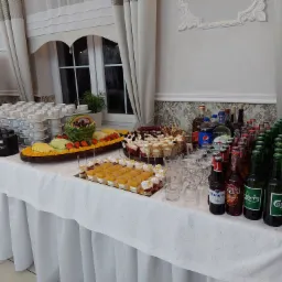 Stół cateringowy z dekoracją z arbuza, paterami owoców, ciastami w pucharkach, napojami w butelkach i kartonikach, oraz stosem filiżanek do kawy, przygotowany na imprezę.