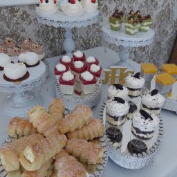 Firma Usługowa Finezja - Bogato zastawiony stół z różnorodnymi deserami: babeczki z owocami, mini torciki, kremowe desery w szklankach z Oreo, kostki galaretki z bitą śmietaną, rurki z kremem posypane cukrem, zielone...