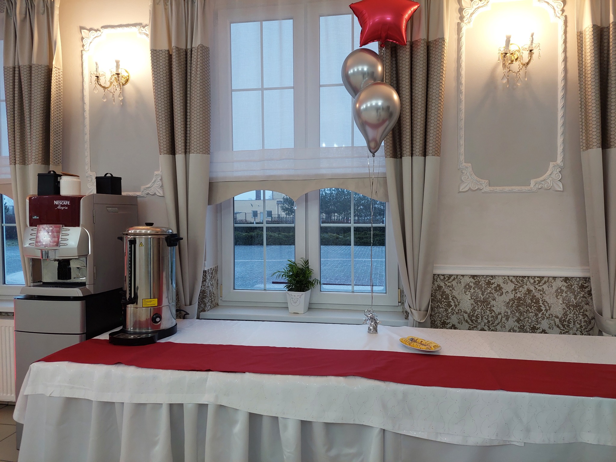 Stół z białym obrusem, czerwonym bieżnikiem, dekoracjami balonowymi, automatem do kawy i podgrzewaczem wody, ustawiony przy oknie z widokiem na zewnątrz, ozdobiony rośliną doniczkową.
