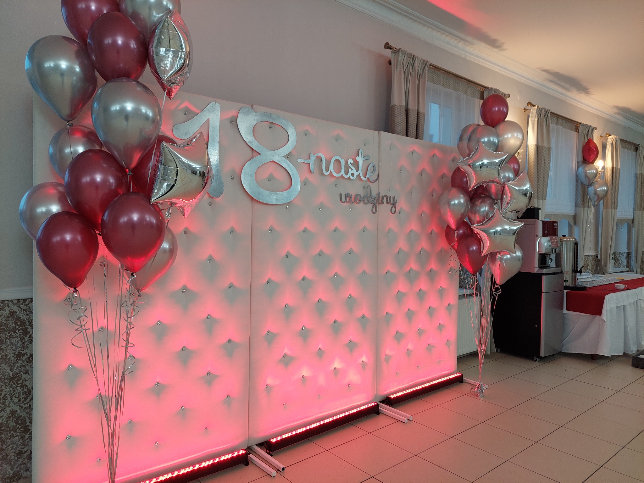 Dekoracja na osiemnaste urodziny: srebrne balony i napis '18-naste urodziny' na białym, pikowanym tle z czerwonym podświetleniem.
