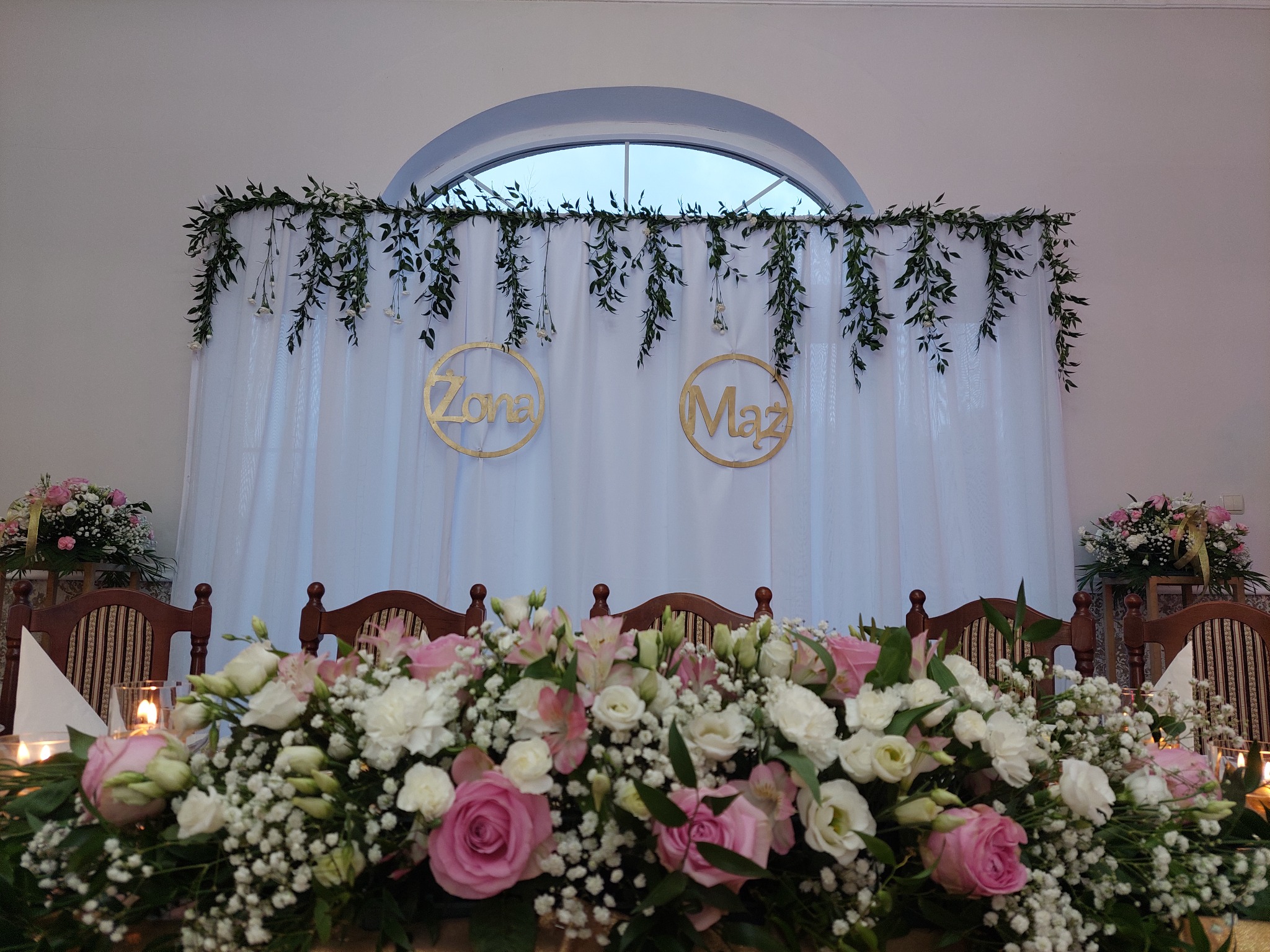 Udekorowany stół weselny z kwiatami, świecami i napisem 'Żona' i 'Mąż' na tle białej zasłony i zielonych girland.