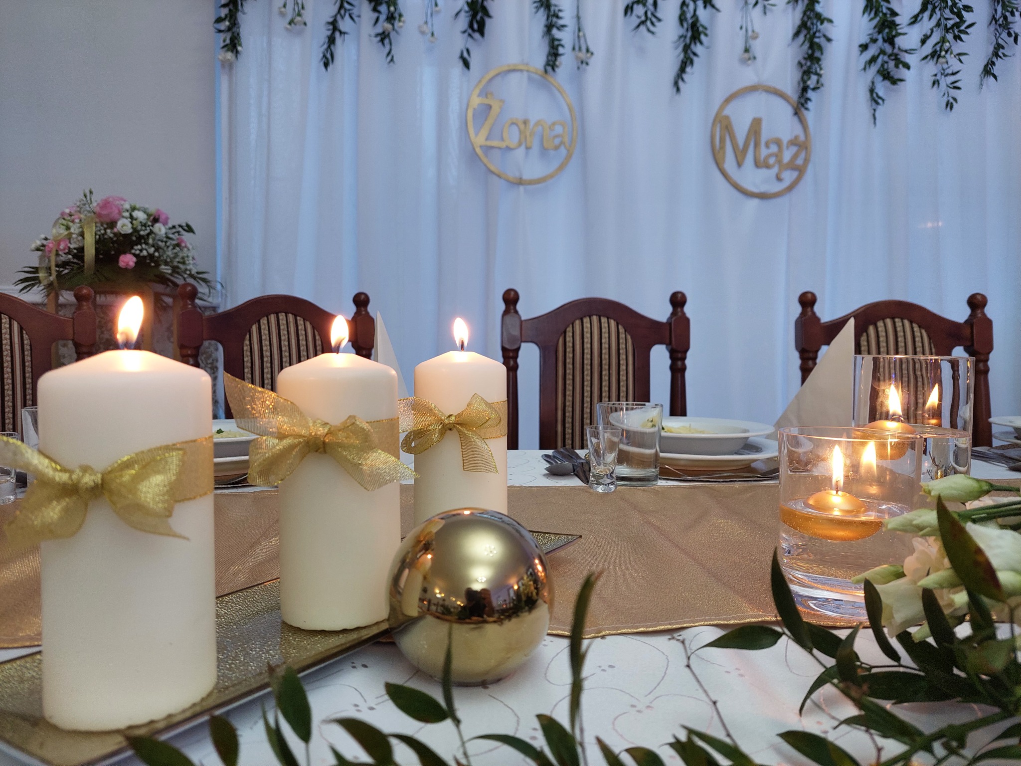 Uroczysty stół z białym obrusem, złotym bieżnikiem, świecami z kokardami, kwiatami i dekoracjami 'Żona' i 'Mąż' na tle białej zasłony.