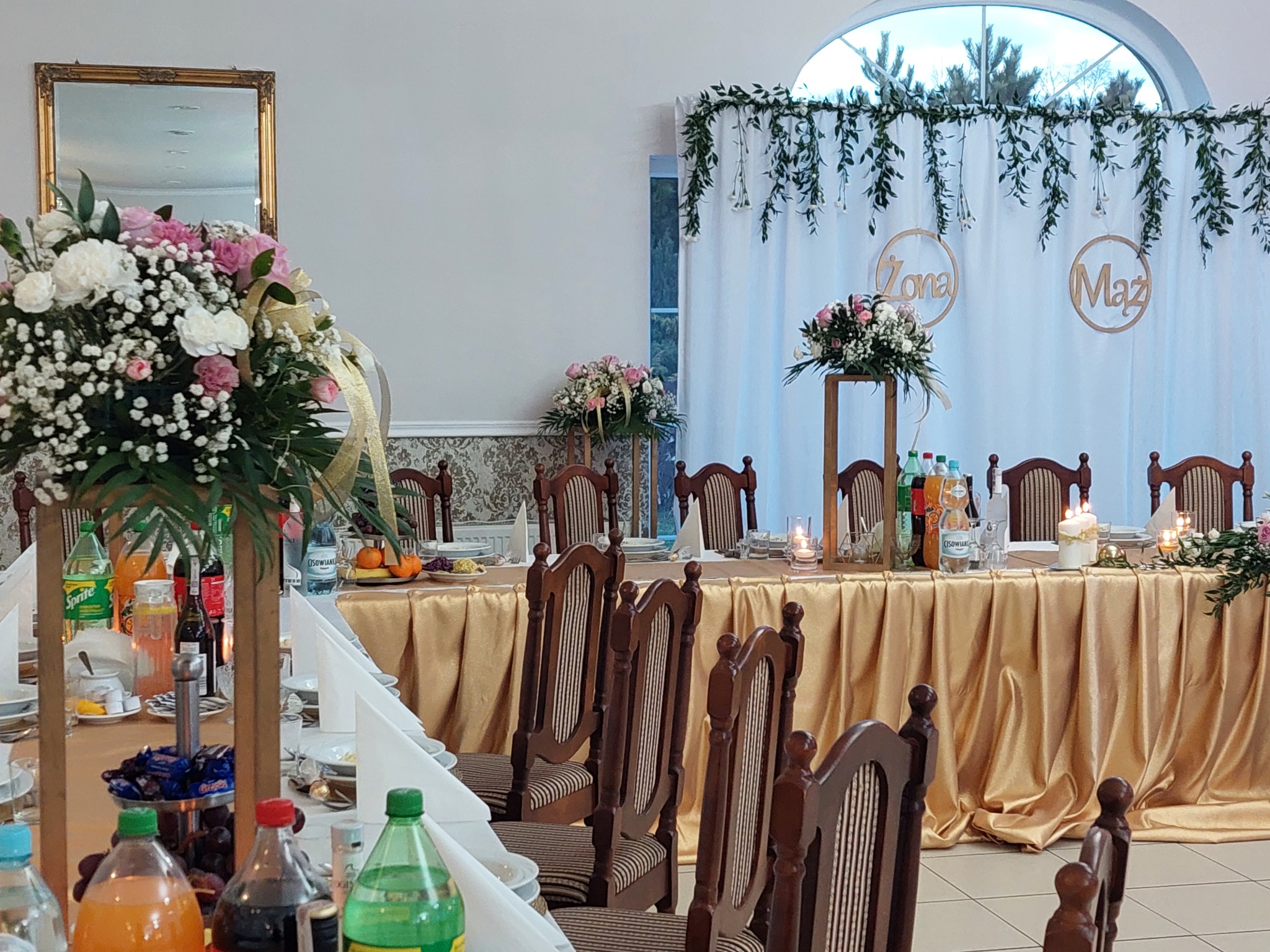 Uroczysty stół weselny z dekoracją kwiatową, złotym obrusem i napojami, tło z napisem 'Żona' i 'Mąż' oraz girlandami z zielonych liści.