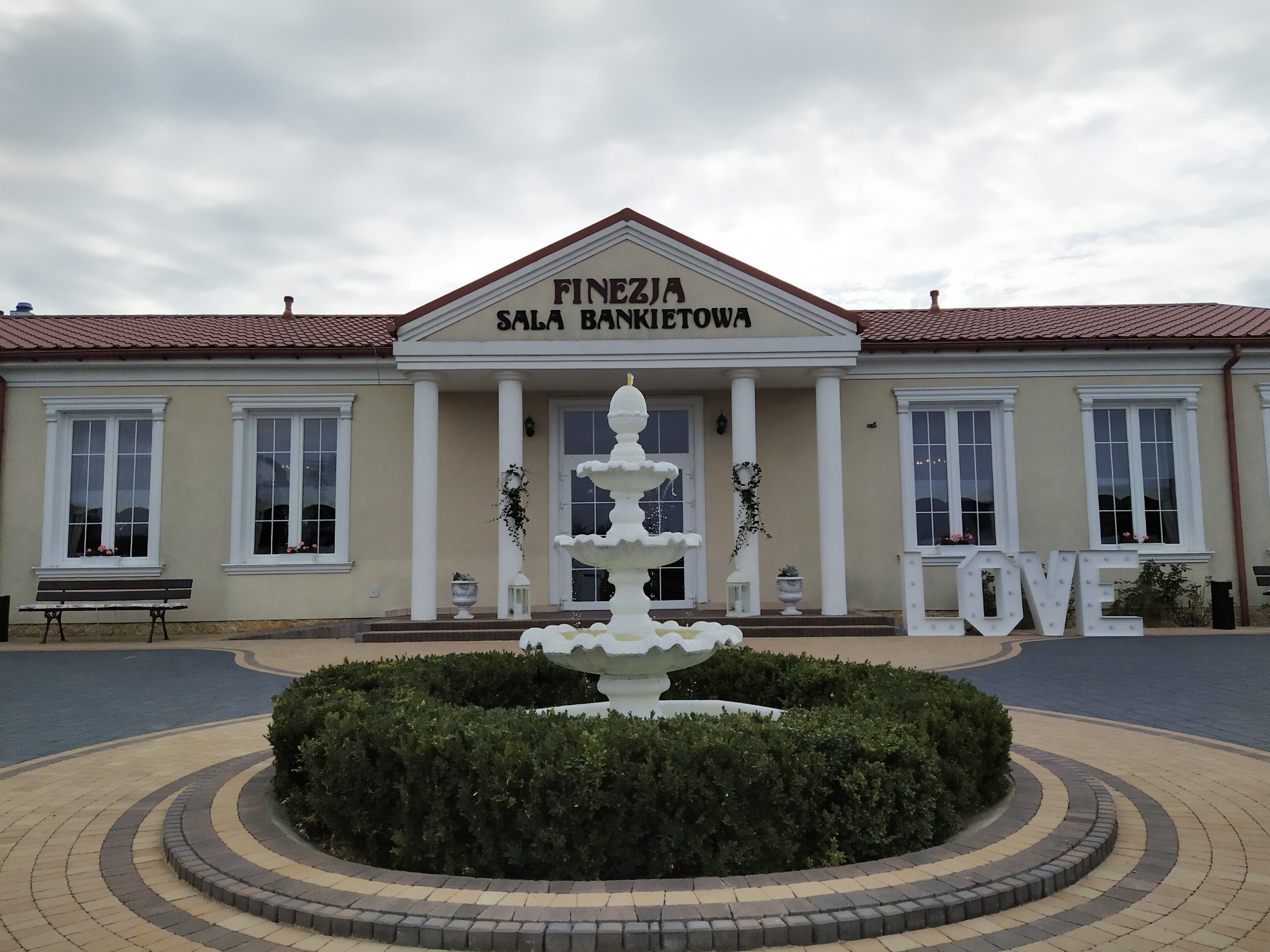 Elewacja budynku sali bankietowej z napisem 'Finezja Sala Bankietowa', fontanna na pierwszym planie, dekoracja 'LOVE' po prawej stronie.