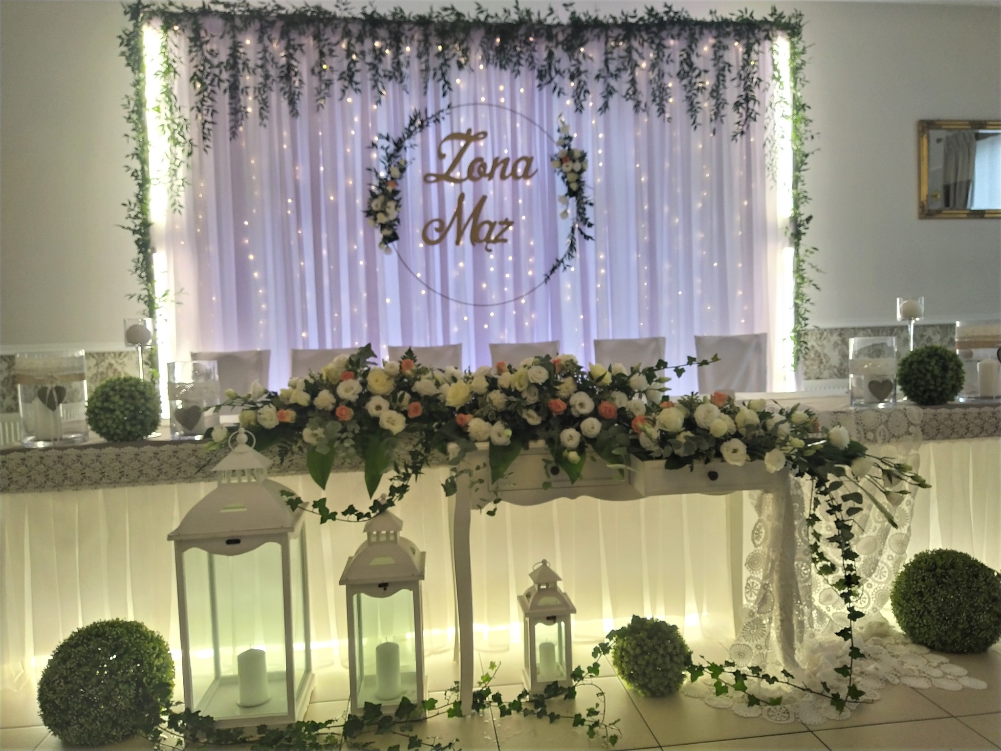 Dekoracja stołu weselnego z białymi lampionami, kompozycją kwiatową z białych i delikatnie różowych kwiatów oraz tłem z białej tkaniny z drobnymi światełkami i napisem 'Żona Mąż' w okręgu...