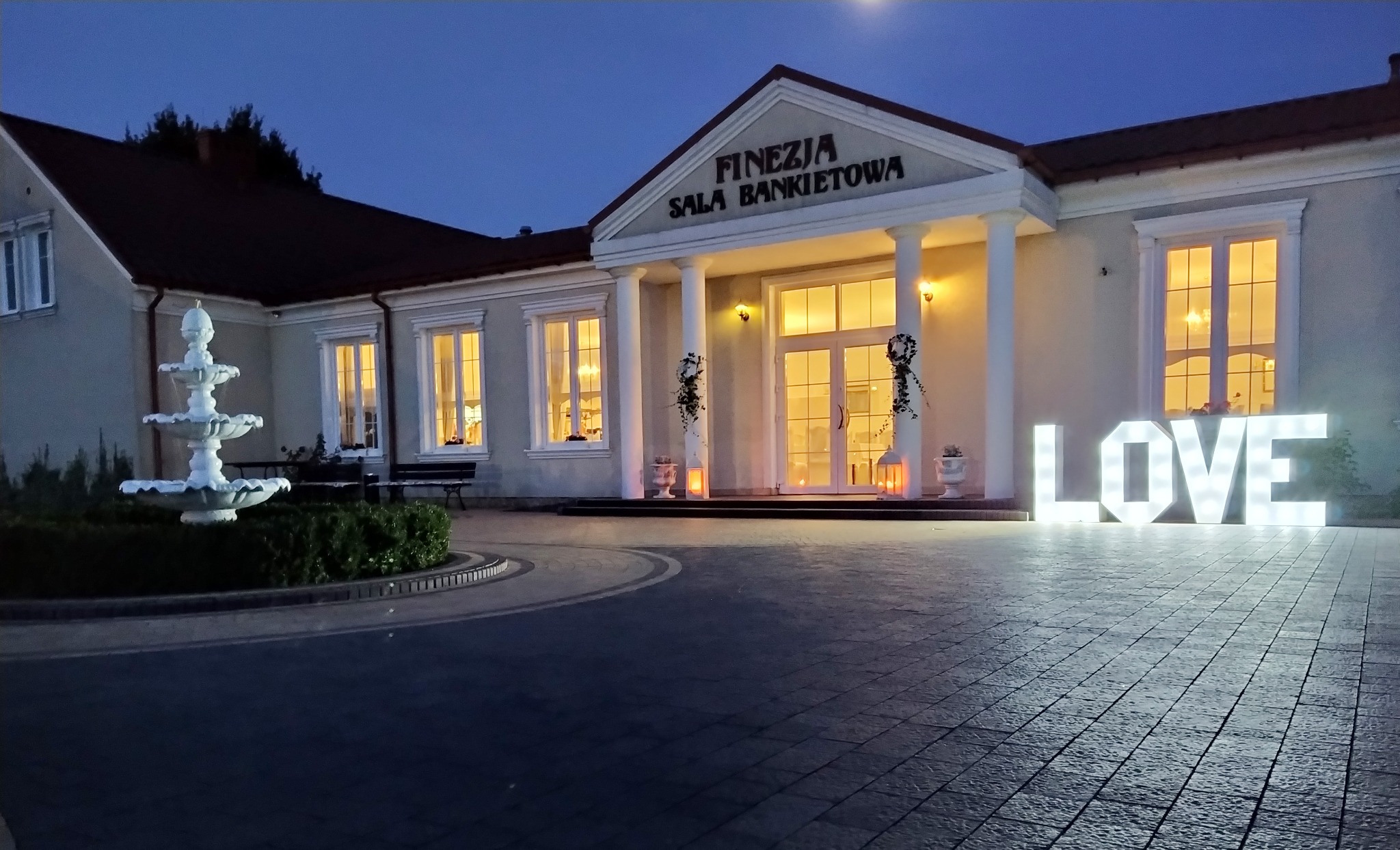 Elewacja sali bankietowej 'Finezja' oświetlona wieczorem, z dekoracyjnym napisem 'LOVE' i fontanną na pierwszym planie.