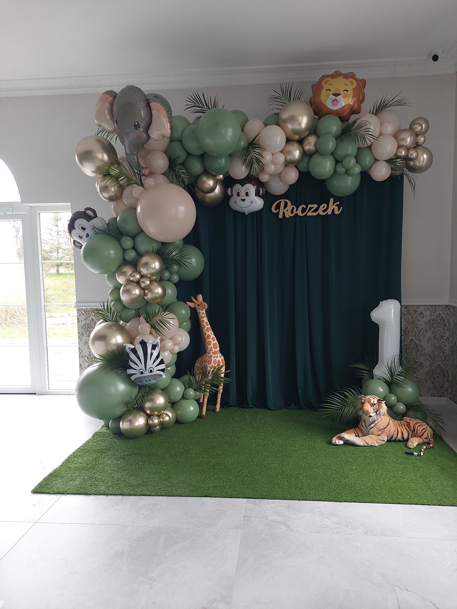 Dekoracja balonowa na roczek w stylu safari z balonami w kształcie zwierząt, zielonymi i złotymi balonami, napisem 'Roczek' i pluszowym tygrysem na sztucznej trawie.