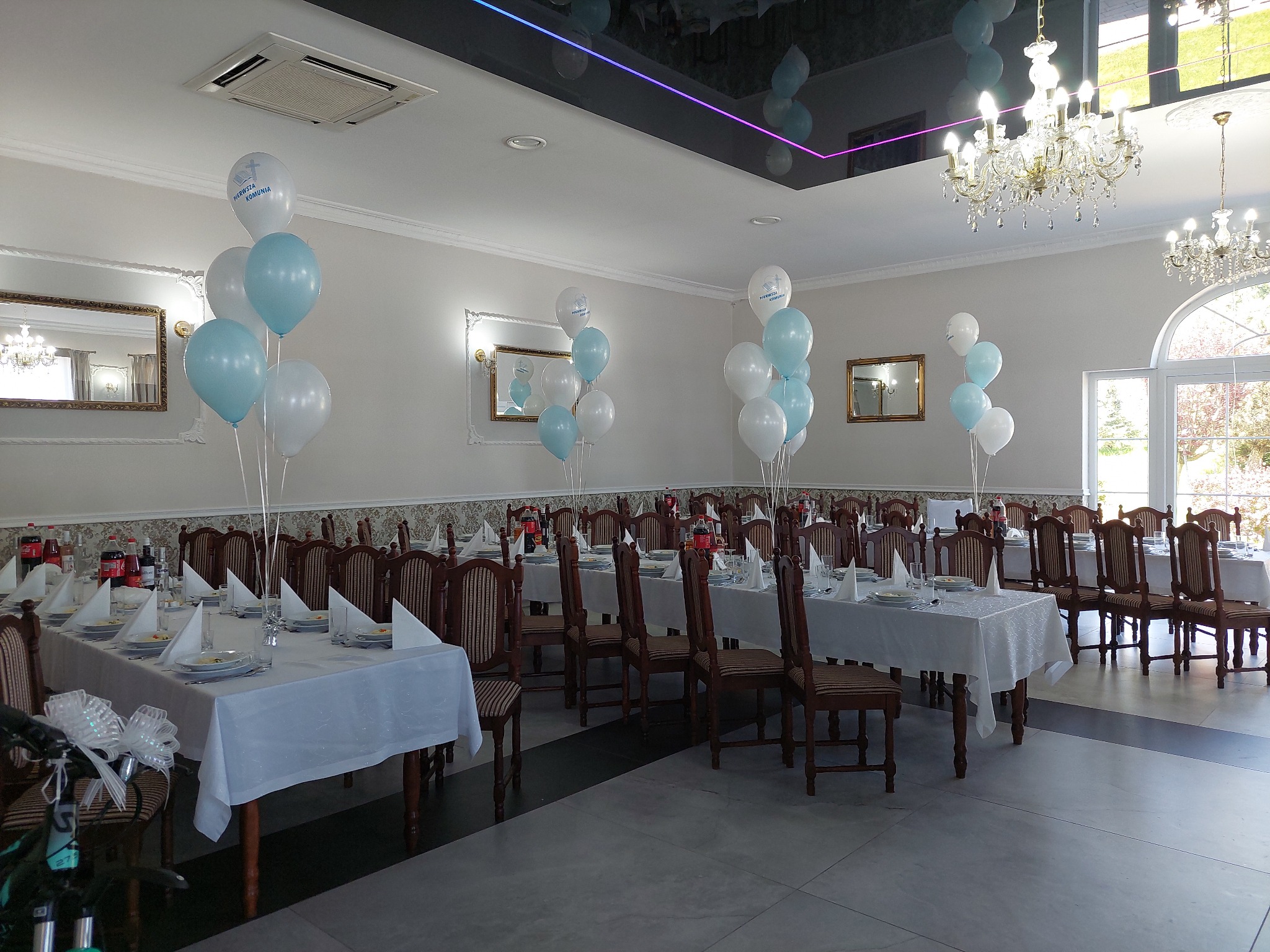 Uroczysta sala z nakrytymi stołami, udekorowana białymi i błękitnymi balonami z nadrukiem komunijnym, kryształowe żyrandole i lustra na ścianach.