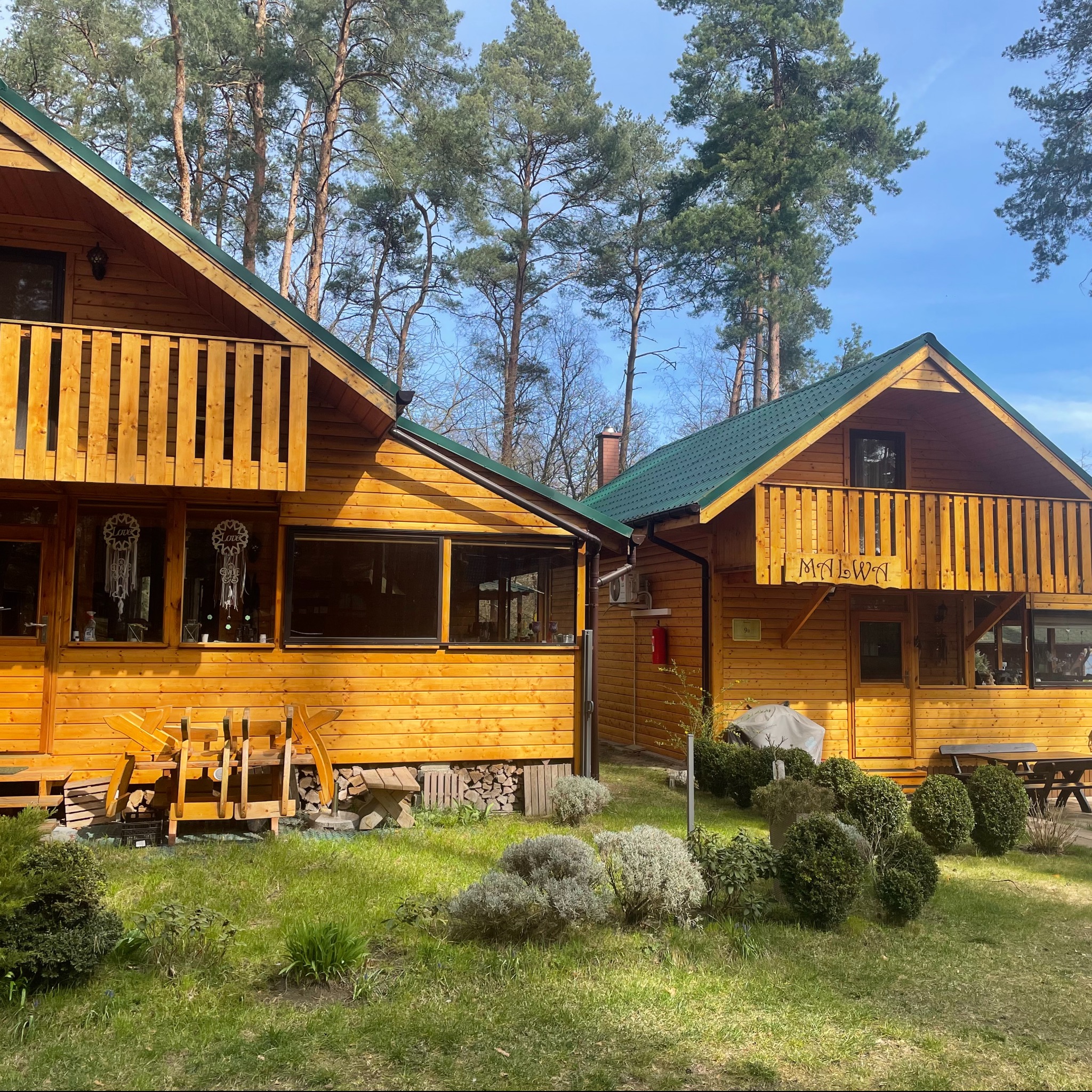 Dwa drewniane domki letniskowe z balkonami, pomalowane na żółto, z zielonymi dachami, otoczone zielenią i drzewami.