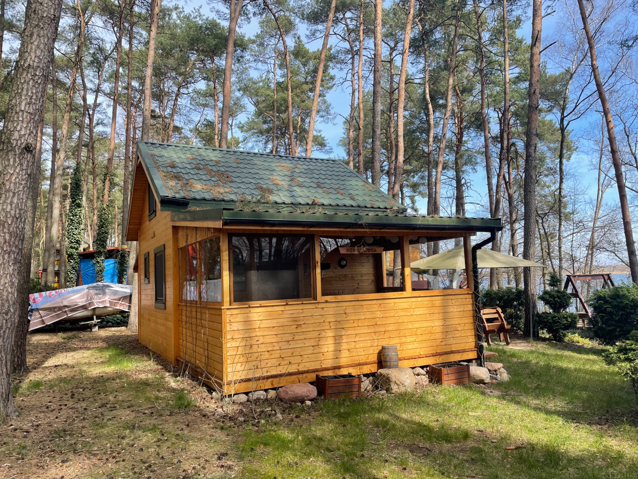 Drewniana altana z zielonym dachem w otoczeniu wysokich sosen, z widocznymi elementami wykończenia zewnętrznego i fragmentem jeziora w tle.