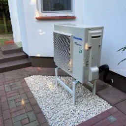 Zewnętrzna jednostka klimatyzacji Panasonic Inverter R32, zamontowana na metalowym stelażu na tle białej ściany domu z oknem i brukowanym podłożem wysypanym białym kamieniem.