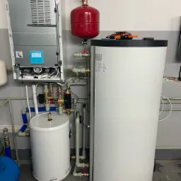 Zestaw urządzeń grzewczych: otwarty panelowy kocioł, czerwony zbiornik ciśnieniowy, biały zasobnik ciepłej wody i duży biały zbiornik akumulacyjny, połączone rurami z zaworami