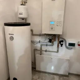 Biały zasobnik ciepłej wody użytkowej Kospel z czarnym deklem, podłączony do pompy ciepła Panasonic Aquarea Smart Cloud, widoczne rury i zawory instalacji w pomieszczeniu technicznym.