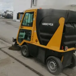 Żółto-czarna zamiatarka Karcher w trakcie pracy, zbierająca zanieczyszczenia z betonowej nawierzchni z kratką odpływową.